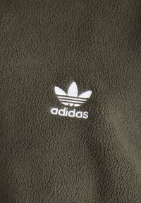 Grön fleecetyg med en vit broderad Adidas-logotyp och tre bladliknande ränder som ger en mjuk och texturerad känsla.