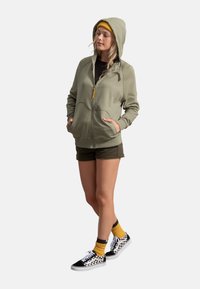 Felpa zip con cappuccio color verde oliva, due tasche frontali, abbinata a pantaloni corti verde oliva, berretto giallo e scarpe da ginnastica a scacchi bianche e nere.
