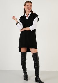 Black knit dress with a V-neck and short cap sleeves, worn over a white collared shirt. Paired with knee-high black leather boots.