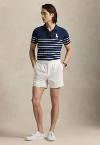 Mężczyzna stojący z rękami w kieszeniach, ubrany w granatową koszulkę polo w paski, białe szorty, białe trampki i okulary przeciwsłoneczne zwisające z kołnierzyka koszulki.