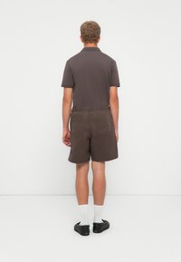 Homme se tenant face à un mur blanc, portant un polo gris foncé, un short marron avec des poches arrière, des chaussettes blanches et des chaussures noires à enfiler.