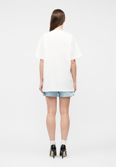 Γυναίκα με μακριά καστανά μαλλιά φοράει λευκό υπερμεγέθες μπλουζάκι, ανοιχτόχρωμα τζιν σορτς, λευκές κάλτσες και μαύρα ψηλοτάκουνα παπούτσια, στέκεται με την πλάτη προς τα εμπρός.