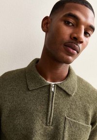 Olivgrön stickad tröja med dragkedja vid halslinningen och en bröstficka. Texturerat tyg och en klassisk krage.