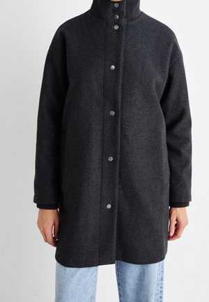 Manteau gris foncé boutonné avec un col montant porté sur un jean bleu clair, visible de la mi-cuisse jusqu'au cou sur un fond uni.