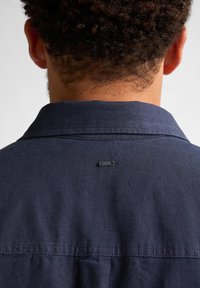 Marinblå skjorta med krage, med en liten logotag på baksidan och ett texturerat tyg. Synlig sömnad längs sömmarna.