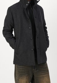 Šedý kabát z vlněné směsi s rybí kostí, s vysokým límcem, zapínáním na knoflíky, detailem s přední kapsou a texturovanou látkou, v kombinaci s tmavými kalhotami.