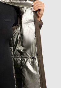 Gepolsterte Jacke mit metallic-silbernem Innenfutter, das eine glatte Textur aufweist, einem schwarzen Panel auf der Außenseite und einem Kordelzugdetail auf der Innenseite.