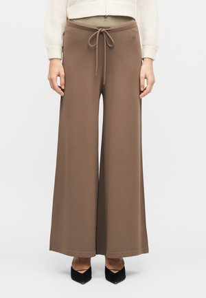 Pantalon marron à jambes larges avec ceinture nouée à l'avant, porté avec des escarpins noirs à bout pointu et un haut crème à manches longues.
