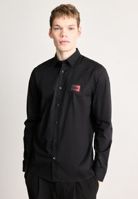 Μαύρο μακρυμάνικο πουκάμισο από λείο ύφασμα, με κουμπιά μπροστά και ορθογώνιο κόκκινο σημάδι με την επιγραφή "HUGO" σε μαύρο κείμενο.