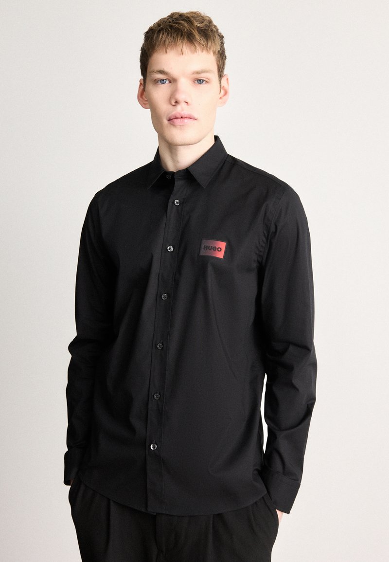 Μαύρο μακρυμάνικο πουκάμισο από λείο ύφασμα, με κουμπιά μπροστά και ορθογώνιο κόκκινο σημάδι με την επιγραφή "HUGO" σε μαύρο κείμενο.