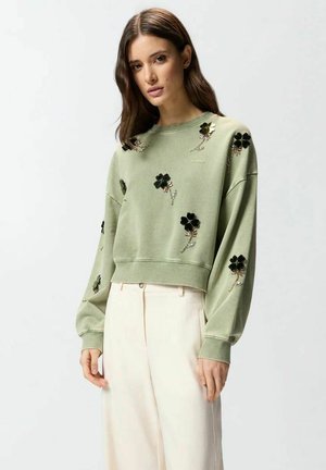 Donna che indossa una felpa verde chiaro con applicazioni floreali nere e pantaloni a vita alta color crema, su uno sfondo chiaro e uniforme.