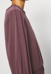 Långärmad klänning i djup mauve, med rund halsringning, samlade puffärmar och en smockad midja för extra textur.