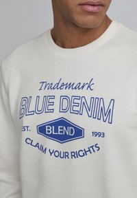 El sudadera blanca presenta texto impreso en azul: "Trademark Blue Denim," con 'Blend' dentro de un hexágono azul, y "Claim Your Rights" debajo.
