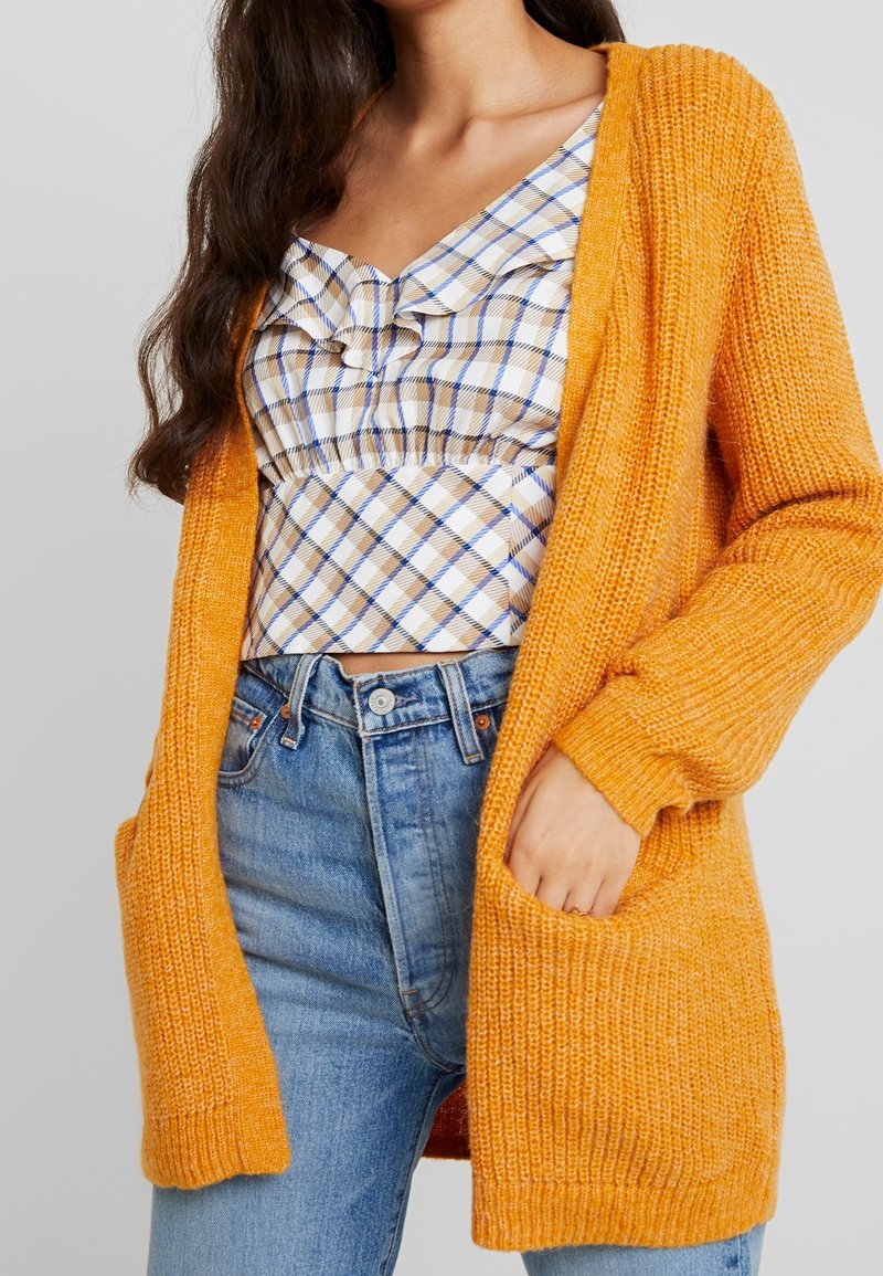 Kvinna i en senapsgul tjockstickad cardigan över en volangig vit och beige rutiga crop-top och högmidjade blå jeanstroppar.