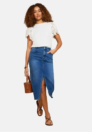 Nainen kiharilla hiuksilla valkoisessa pitsihiha-puserossa, sinisessä farkkujen midi-mekossa, jossa edessä halkio, ruskeat sandaalit ja ruskea käsilaukku kädessään.