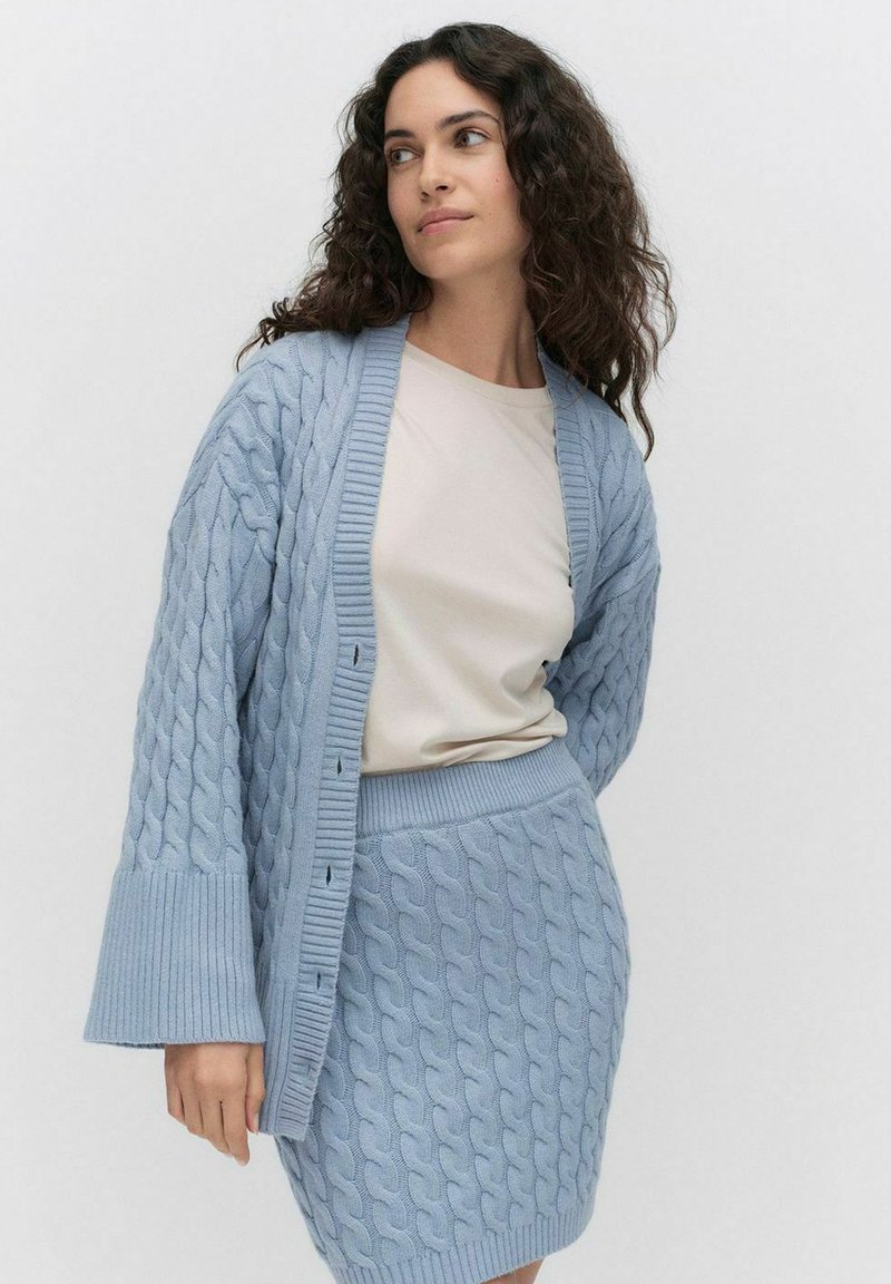 Vaaleansininen neulottu neuletakki, jossa on palmikkokuvio, ylisuurta kokoa, ribbireunat, yhdistettynä yhteensopivaan hameeseen. Sileä pinta, rento muotoilu.