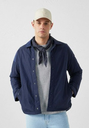 Navy blue lightweight jacket with snap buttons, worn over a gray shirt. Features a dark scarf and a beige cap. Textured fabric with a relaxed fit.
