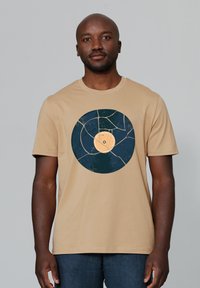 Beige T-shirt met een grafisch ontwerp van een vintage vinylplaat in donkerblauw en oranje, met een gestructureerd cirkelvormig ontwerp en korte mouwen.