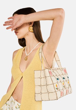 Borsa ricamata con un motivo a griglia in crema, decorata con motivi floreali colorati. Presenta dettagli in perle e robuste maniglie.