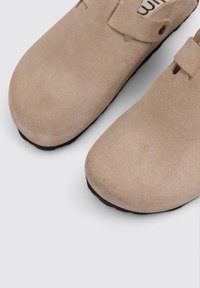 Sabots en suède beige avec une pointe arrondie et une semelle en caoutchouc noire. Dotés d'une sangle réglable et de petites découpes près de la sangle.