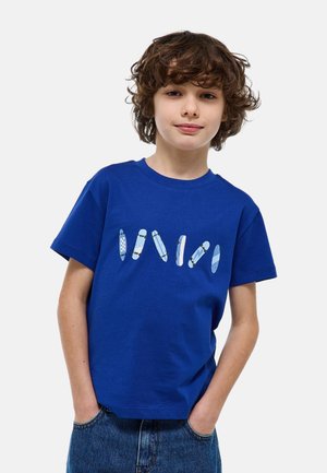 Garçon aux cheveux bouclés bruns portant un t-shirt bleu avec six skateboards illustrés, debout les mains dans les poches de son jean.