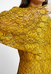 Vestido de encaje con mangas largas en un vibrante color amarillo. Patrones florales intrincados con acentos de hilo negro y bordes flecos.