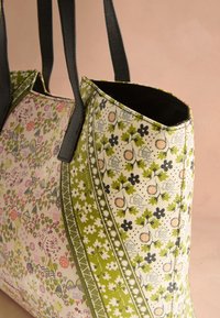 Torba na ramię z czarnymi uchwytami, ozdobiona zielonymi i różowymi kwiatowymi oraz inspirowanymi naturą wzorami na beżowym tle materiału.