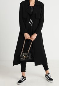 Osoba w czarnym długim płaszczu, czarnych spodniach, topie w kropki, czarno-białych sneakersach, trzymająca czarną torebkę z złotymi ćwiekami i łańcuchowymi ramiączkami.