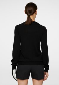 Svart långärmad tröja med ribbade cuffs och nederkant, tillverkad av ett mjukt material, bärs över mörka shorts, ses från baksidan.