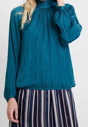 Kvinde iført en tealfarvet bluse med lange ærmer, rynket halsudskæring og elastisk kant, kombineret med et navyblåt stribet nederdel.