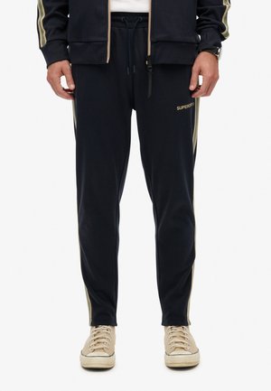 Marineblauwe sweatpants met contrasterende gouden zijstrepen, boord met trekkoord en subtiele branding op de linker bovenbeen. Katoenmix, zachte textuur.