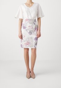 Blouse blanche avec des manches volantées, associée à une jupe ajustée, longueur genou, arborant un motif floral dans des tons violets et roses. Texture claire et lisse.