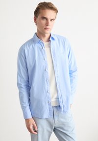 Camicia azzurro chiaro con bottoni in un taglio rilassato, realizzata in tessuto morbido con dettaglio della tasca. Il modello indossa una maglietta bianca sotto.