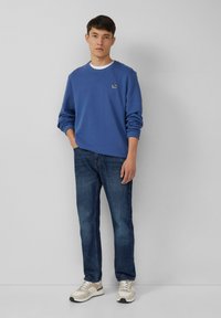 Giovane uomo che indossa una felpa blu, jeans scuri e sneakers bianche, in piedi con una mano in tasca contro uno sfondo grigio semplice.