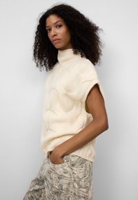 Kremowy sweter dziany z wysokim dekoltem i krótkimi rękawami, z teksturowanym wzorem. W połączeniu z jasnymi spodniami w marmurkowy wzór.