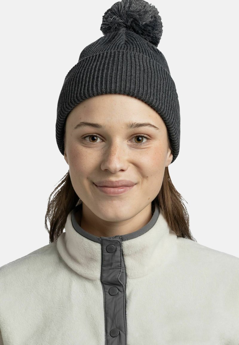 Gray knitted beanie with a pom-pom on top, featuring a ribbed texture. Worn over a cream fleece jacket with gray button detailing.