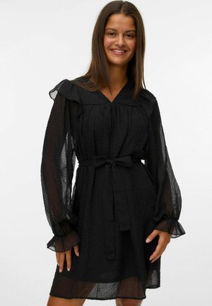 Femme souriante, portant une robe noire à manches longues avec des manches en voile, des poignets à volants et une ceinture nouée à la taille, se tenant devant un fond uni.