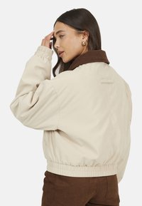 Veste bomber beige avec un col en velours côtelé marron, des poignets et une taille élastiques, présentant une texture simple et lisse ainsi qu'un design minimaliste.