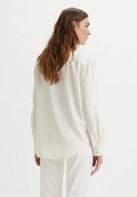 Camisa blanca de manga larga con un corte relajado, que presenta puños abotonados, textura suave y un dobladillo curvado en la parte trasera.