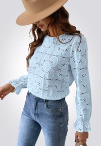 Blusa de encaje azul claro con patrones florales, mangas largas con puños con volantes, combinada con vaqueros oscuros ajustados. Los accesorios incluyen un sombrero.