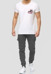T-shirt bianco in cotone con una grafica di una palma rosa e nera sul taschino sinistro, abbinata a jogging grigi in cotone e sneaker bianche.