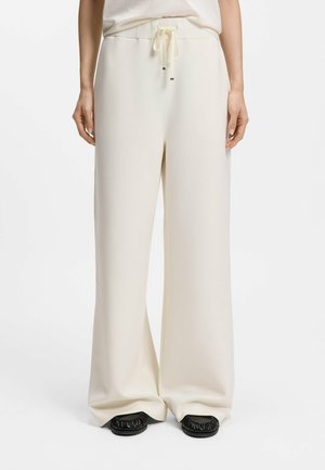 Personne portant un pantalon ample couleur crème à jambes larges avec une taille à cordon et des chaussures noires sans lacets, debout devant un fond blanc.