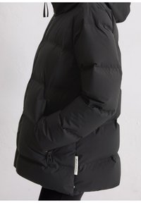 Schwarze gepolsterte Winterjacke mit Kapuze, seitlicher Reißverschluss-Tasche und Hand teilweise in der Tasche, Etikett sichtbar in der Nähe des Saums.