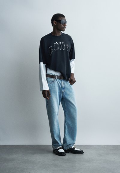 Άνδρας που φοράει υπερμεγέθη μαύρο πουκάμισο πάνω από λευκά μακριά μανίκια, φαρδιά ανοιχτό μπλε τζιν, ασπρόμαυρα παπούτσια και μεγάλα μαύρα γυαλιά ηλίου, στέκεται μπροστά σε γκρι τοίχο.