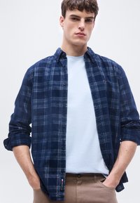 Νέος άνδρας που φοράει πουκάμισο με κουμπιά σε ναυτικό μπλε καρό πάνω από λευκό t-shirt με μπεζ παντελόνι, τα χέρια στις τσέπες, με φόντο απλό.