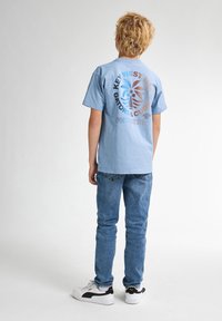 Světle modré bavlněné tričko s kruhovou grafikou, která zobrazuje text a stylizovaný design palmy na zadní straně. Kombinováno s modrými džíny a teniskami.