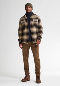 Bruine en beige geruite overhemdjas met borstzakken, gedragen over een zwarte coltrui, gecombineerd met beige broek en bruine veterschoenen. Olijfkleurige beanie.