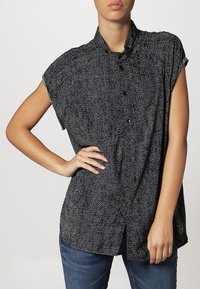 Chemise noire à manches courtes avec un motif tacheté blanc, fermeture à boutons et col. Portée avec un jean bleu.