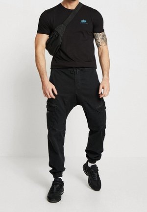 Maglietta nera con logo blu, pantaloni cargo neri con tasche con cerniera, scarpe da ginnastica nere e una borsa a tracolla nera. Vestibilità e design casual.