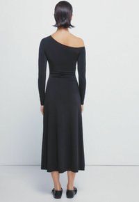 Zwarte jurk met lange mouwen, één schouder bloot, aansluitend bovenlijf en een flowy rok. Glad materiaal en enkellange stijl.